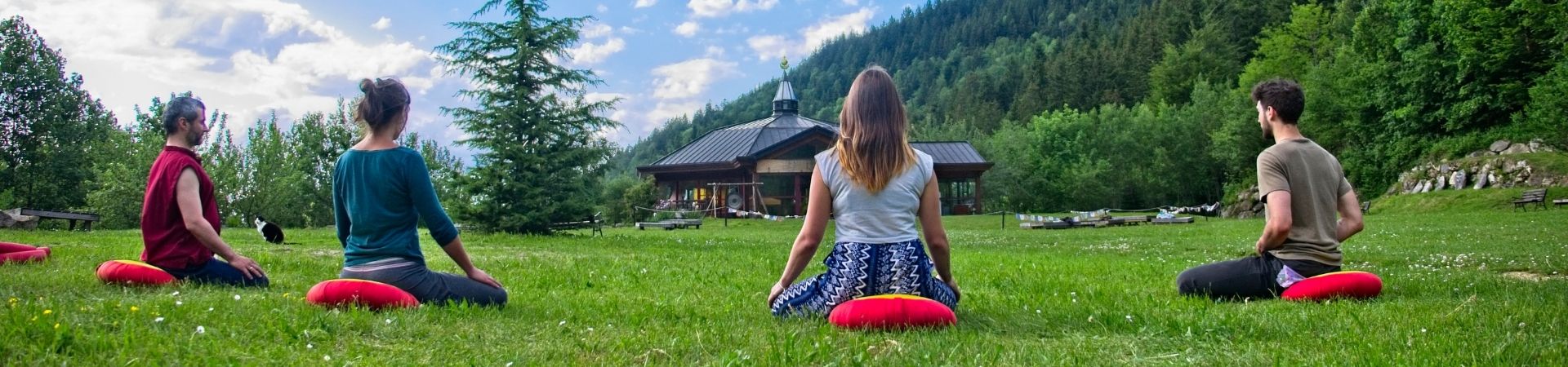 Quatre pratiquants en méditation assise sur coussins rouges dans le jardin de Nigu Karma Ling face au temple, entourés de nature